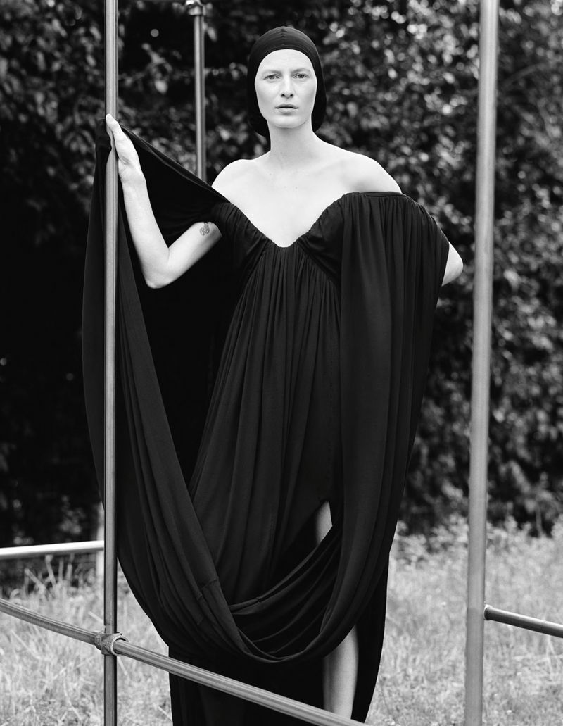 Monochrome portrait of a woman in a flowing black dress posing outdoors with an architectural metal frame element, with lush foliage backdrop.