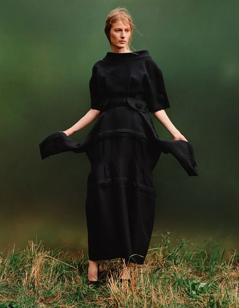 A poised woman in a striking black dress stands in a grassy field against a muted green backdrop, captured in a medium shot.