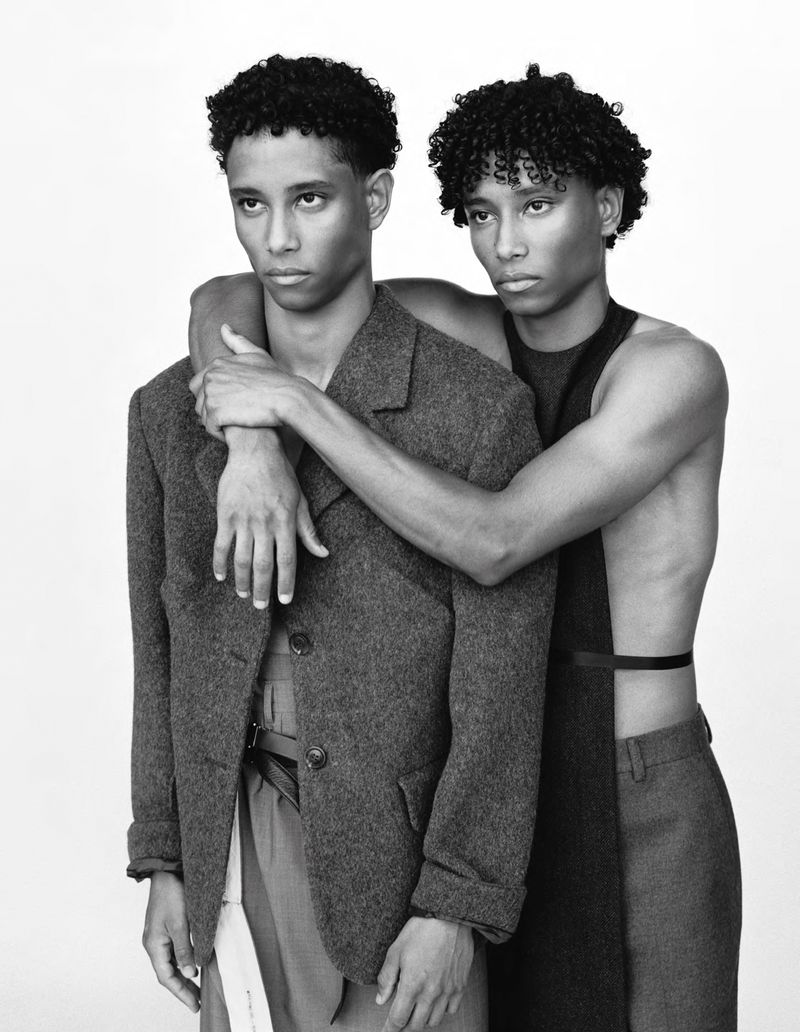 Black and white portrait of two young men in stylish jackets and tops, embracing in a studio setting against a neutral backdrop.