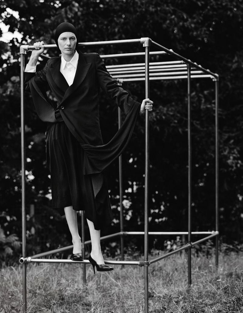 Woman in black suit poses inside metal structure in black-and-white outdoor fashion shot with trees in background. Editorial portrait.