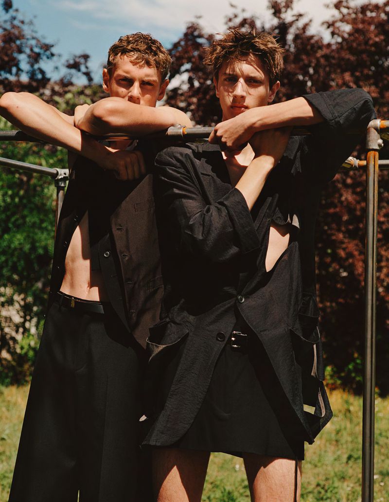 Two young adult male fashion models pose outdoors wearing black linen open button-down shirts and black bottoms near metal pipes.