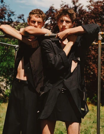 Two young adult male fashion models pose outdoors wearing black linen open button-down shirts and black bottoms near metal pipes.