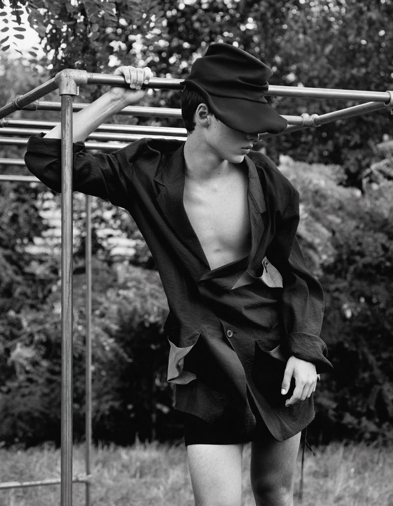 A young man in a dark jacket and hat poses near metal bars in a black and white outdoor shot, blending fashion and natural light.