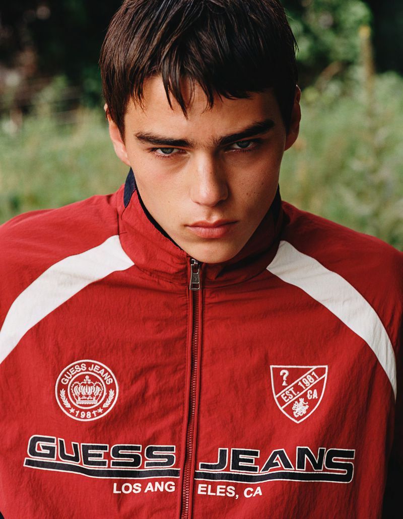 Young man with dark hair poses outdoors wearing a red and white Guess Jeans track jacket against a blurred green background.