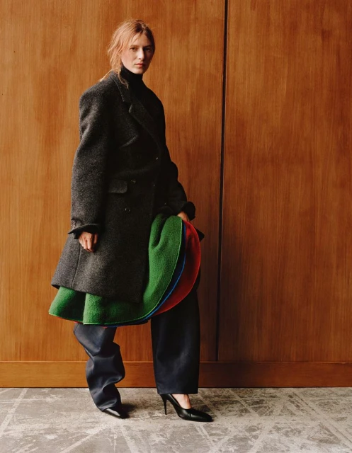 Fashionable woman in a dark coat and wide-leg pants poses against a wood paneled wall, holding a colorful garment. High fashion portrait.