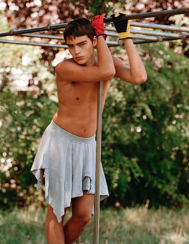 A person in a gray skirt and work gloves poses outdoors, leaning against a pole, with dappled sunlight filtering through the trees.