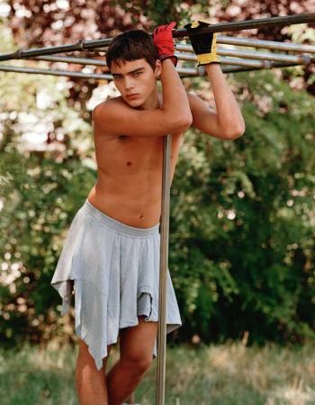 A person in a gray skirt and work gloves poses outdoors, leaning against a pole, with dappled sunlight filtering through the trees.