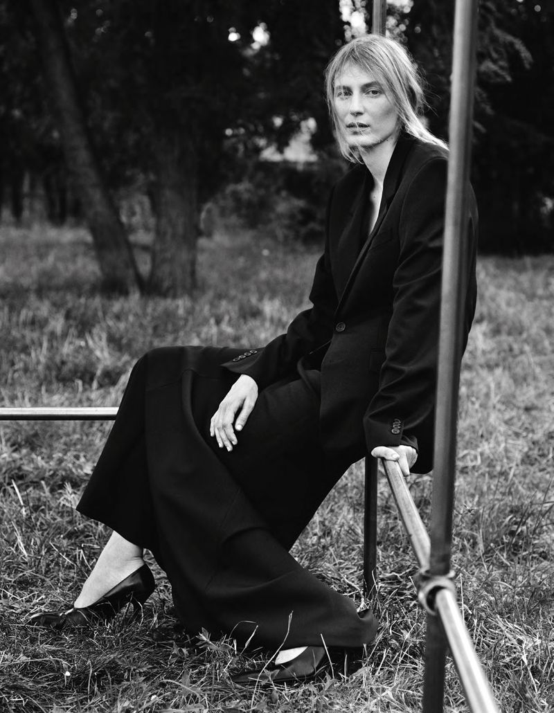 Model wearing a dark tailored suit and skirt seated outdoors near a metal frame structure in a monochrome portrait.