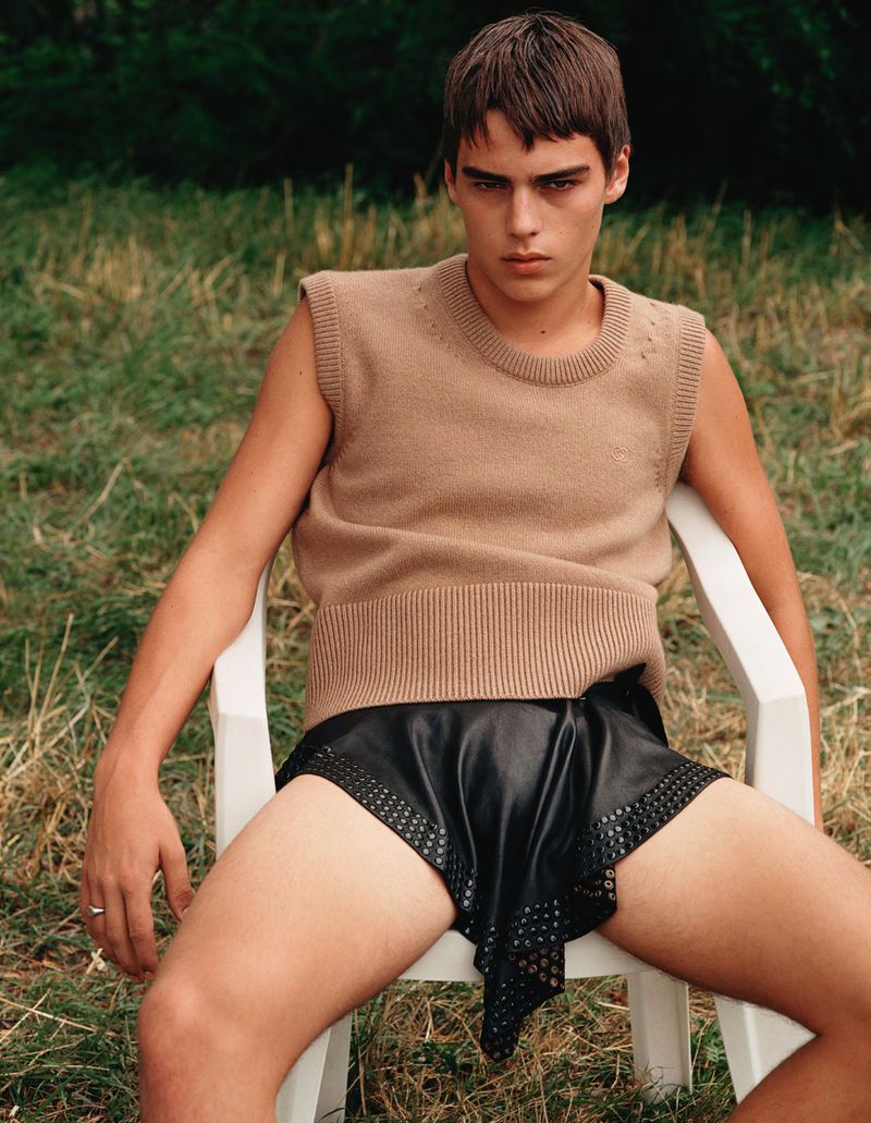 A young adult male poses in a sleeveless knit top and black shorts, seated on a white plastic chair in an outdoor grassy environment.