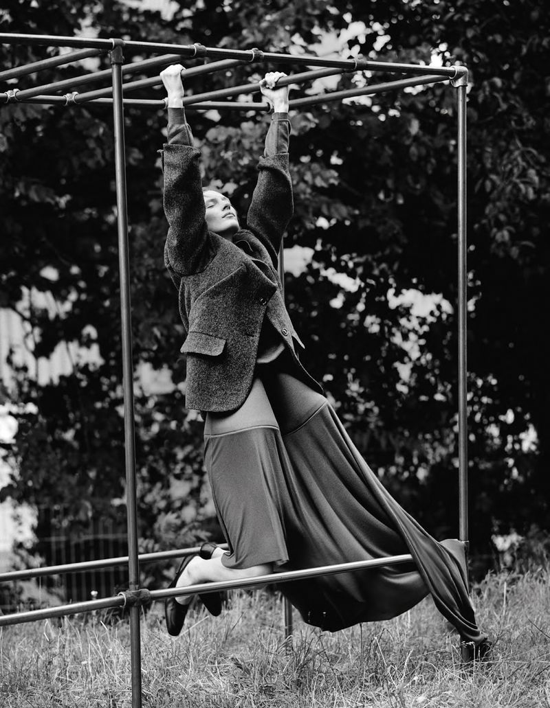 A woman in a grey jacket and long skirt hangs from metal playground bars in a black and white outdoor fashion editorial shot.