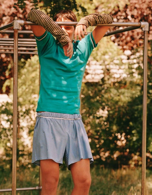 A young man leans on a metal bar structure outdoors, wearing a turquoise shirt and blue striped shorts, with trees blurred in the background.
