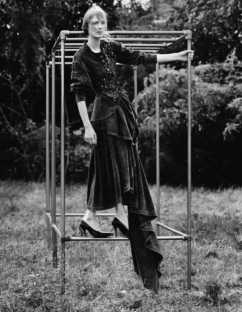 A woman in an elaborate black dress stands on a jungle gym in a grassy outdoor setting, captured in a black and white editorial style.