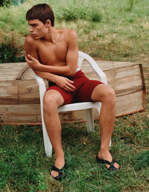 Young man sits on a white plastic chair outdoors in a backyard with grass and a wooden structure. He wears maroon shorts and black sandals.