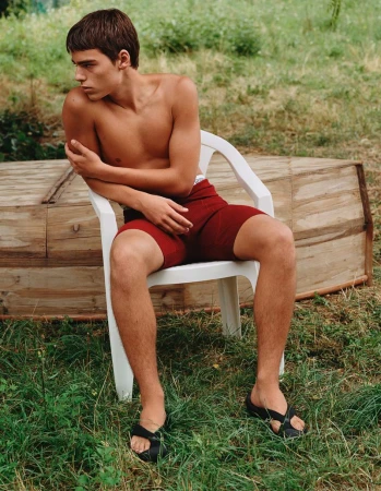 Young man sits on a white plastic chair outdoors in a backyard with grass and a wooden structure. He wears maroon shorts and black sandals.