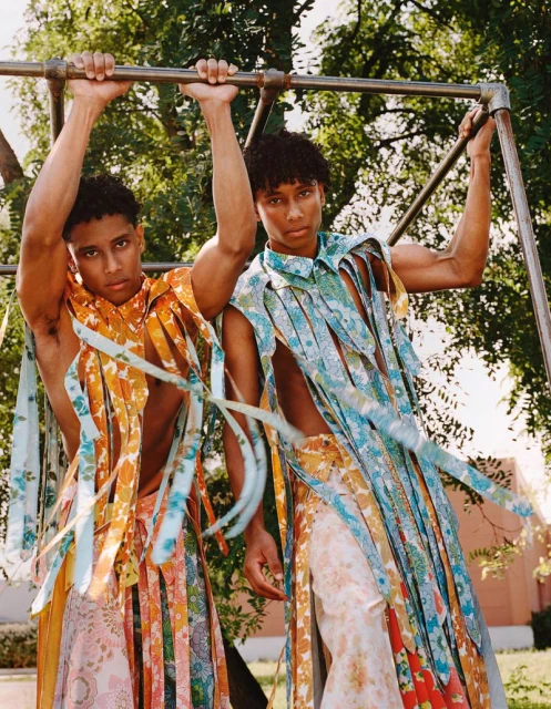 Two men pose in floral-patterned clothing, one grasping a metal bar outdoors with a building and green foliage in the background.