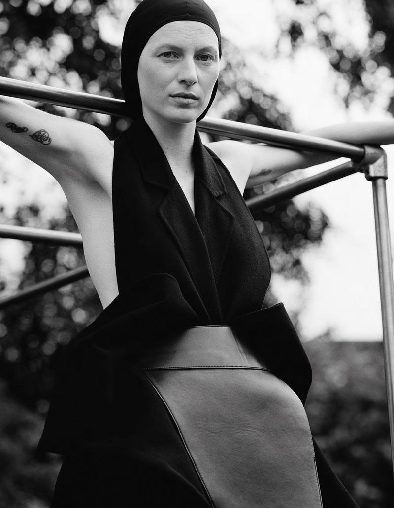 Monochrome portrait of a fair-skinned woman in a hood and black dress posing outdoors with a metal bar structure behind her.