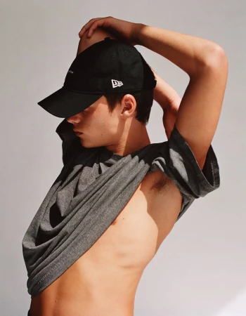 A male fashion model poses against a neutral backdrop, wearing a New Era cap and a gray T-shirt pulled down to reveal his torso.