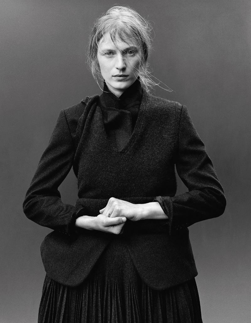 Black and white portrait of a woman in a dark dress and blazer, posing with hands clasped, against a neutral gray background in studio lighting.