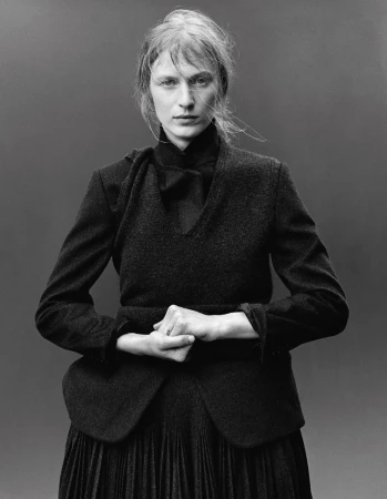 Black and white portrait of a woman in a dark dress and blazer, posing with hands clasped, against a neutral gray background in studio lighting.