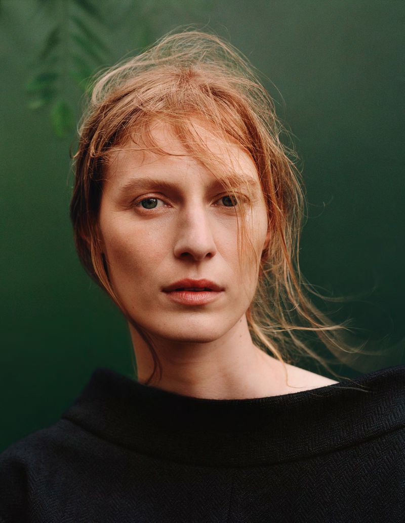 Portrait of a light-skinned adult female with reddish blonde hair, wearing a black top, posing against a blurred green background with leaves.