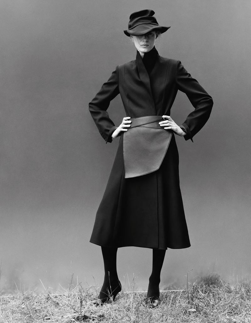 Model wearing a black structured coat, wrap skirt, and draped hat against a grey backdrop.