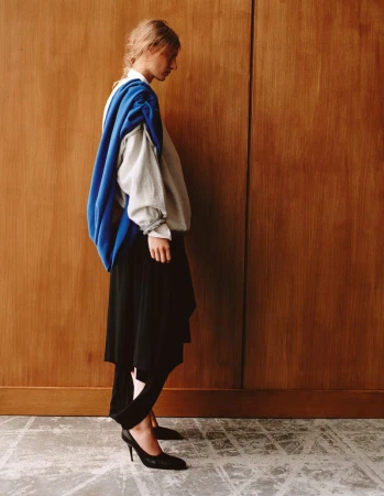 Model Julia Nobis standing in profile wearing a draped blue wrap, grey sweater, black skirt, and heels against a wood wall.