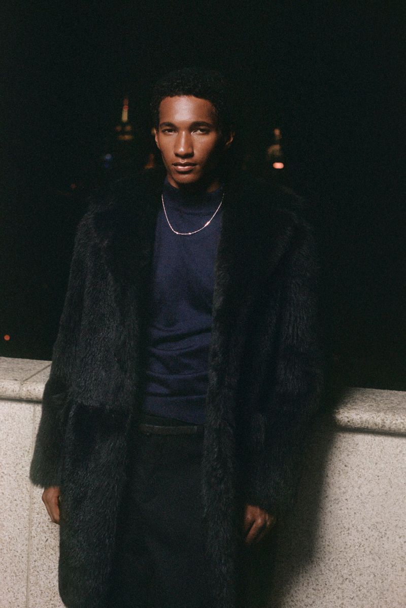 Portrait of an adult male with dark hair wearing a fur coat and navy shirt, photographed outdoors at night with city lights in the background.