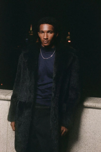 Portrait of an adult male with dark hair wearing a fur coat and navy shirt, photographed outdoors at night with city lights in the background.