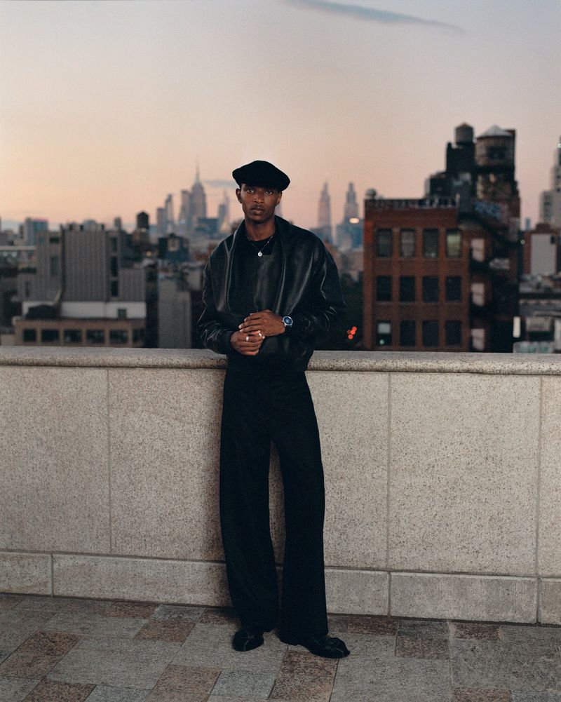 Portrait of stylish adult male in black leather jacket and beret, posed on a rooftop with a cityscape at sunset.