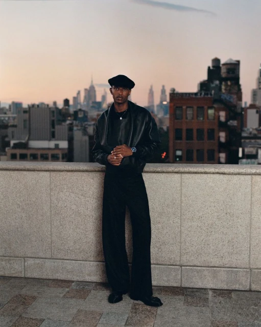 Portrait of stylish adult male in black leather jacket and beret, posed on a rooftop with a cityscape at sunset.