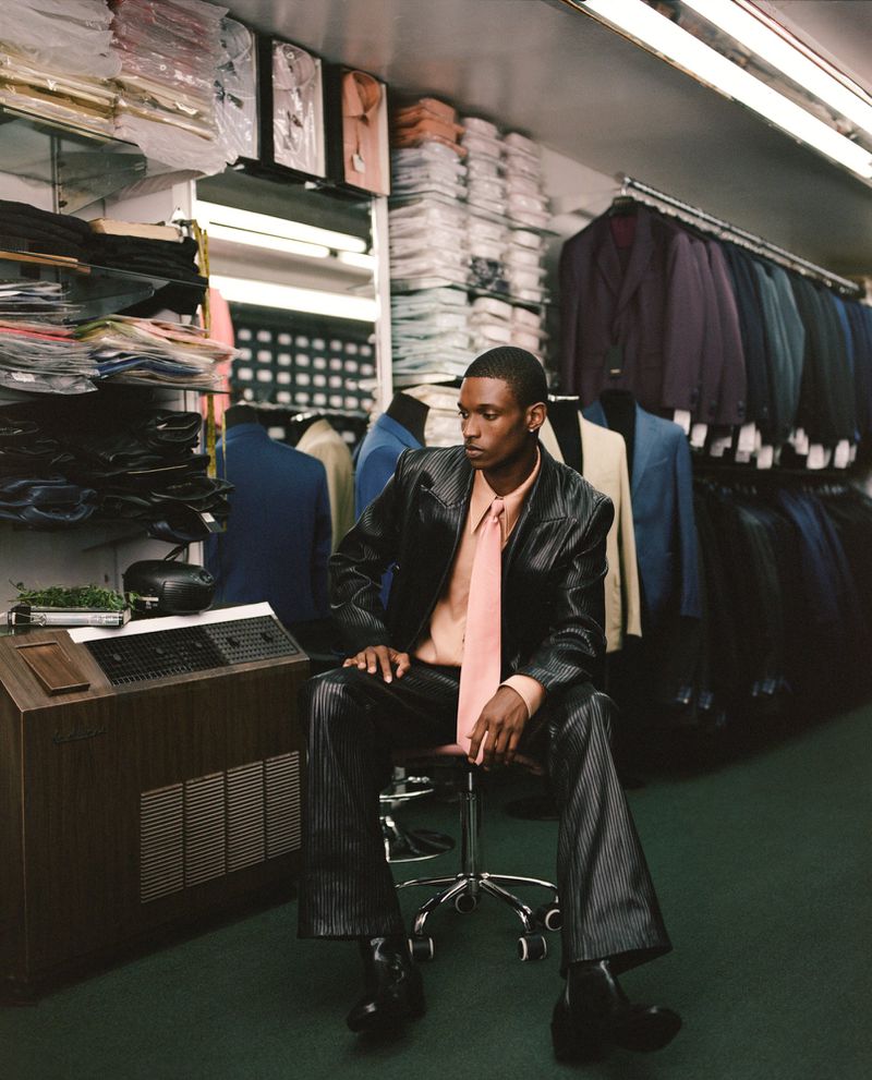 An African-American man in a suit sits in a tailor shop, surrounded by suits and shirts on shelves and racks, with bright overhead lighting.
