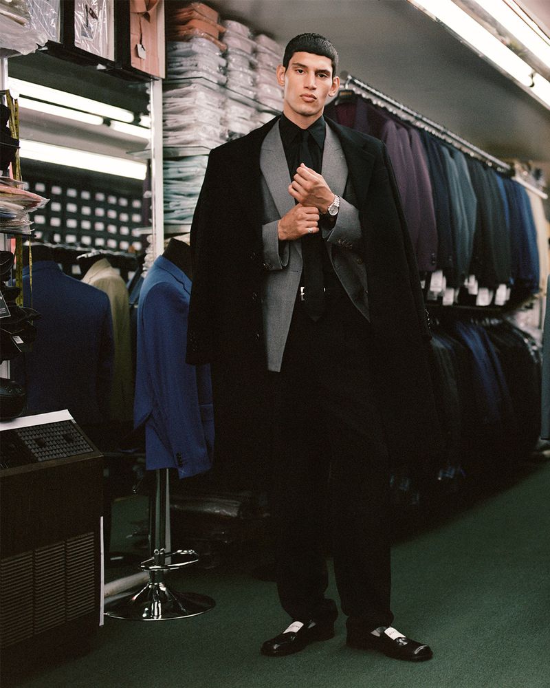 Man in suit poses in a tailor shop. Man wears a black coat over a grey suit, black shirt, black tie, and black loafers with white socks.