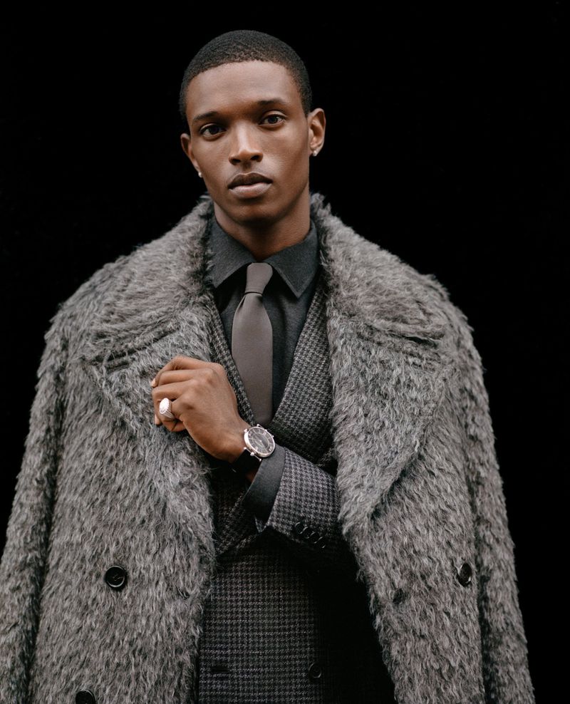 An African-American male model poses in an indoor setting, wearing a grey suit, tie, overcoat and wristwatch against a dark background.