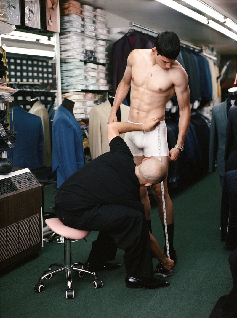 Shirtless man getting measured by tailor inside a clothing store with suits and garments in the background. Fitting session indoors.