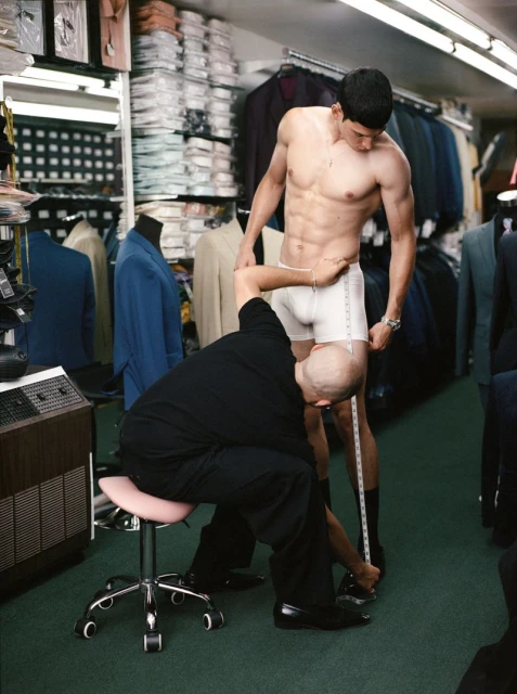 Shirtless man getting measured by tailor inside a clothing store with suits and garments in the background. Fitting session indoors.