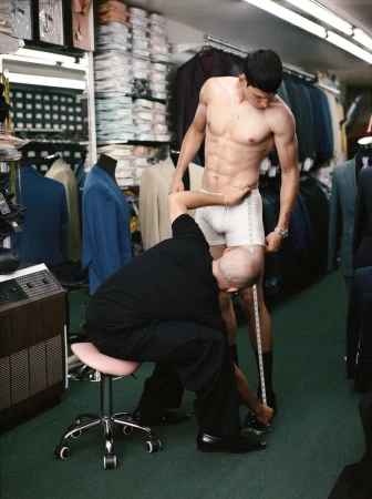 Shirtless man getting measured by tailor inside a clothing store with suits and garments in the background. Fitting session indoors.