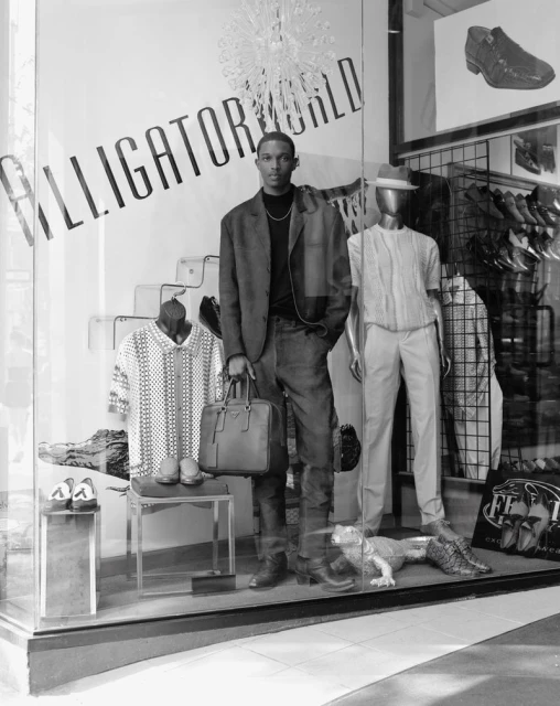 An African-American male model stands in front of a shop window wearing a blazer and jeans next to a mannequin and clothing display.