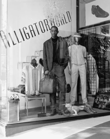 An African-American male model stands in front of a shop window wearing a blazer and jeans next to a mannequin and clothing display.