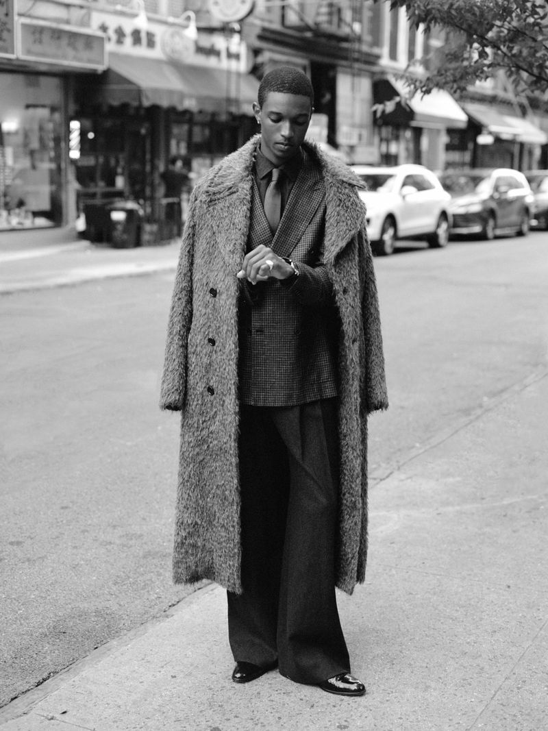 Monochrome portrait of a Black man wearing a brown overcoat over a suit, standing on a city street and looking at his wristwatch.