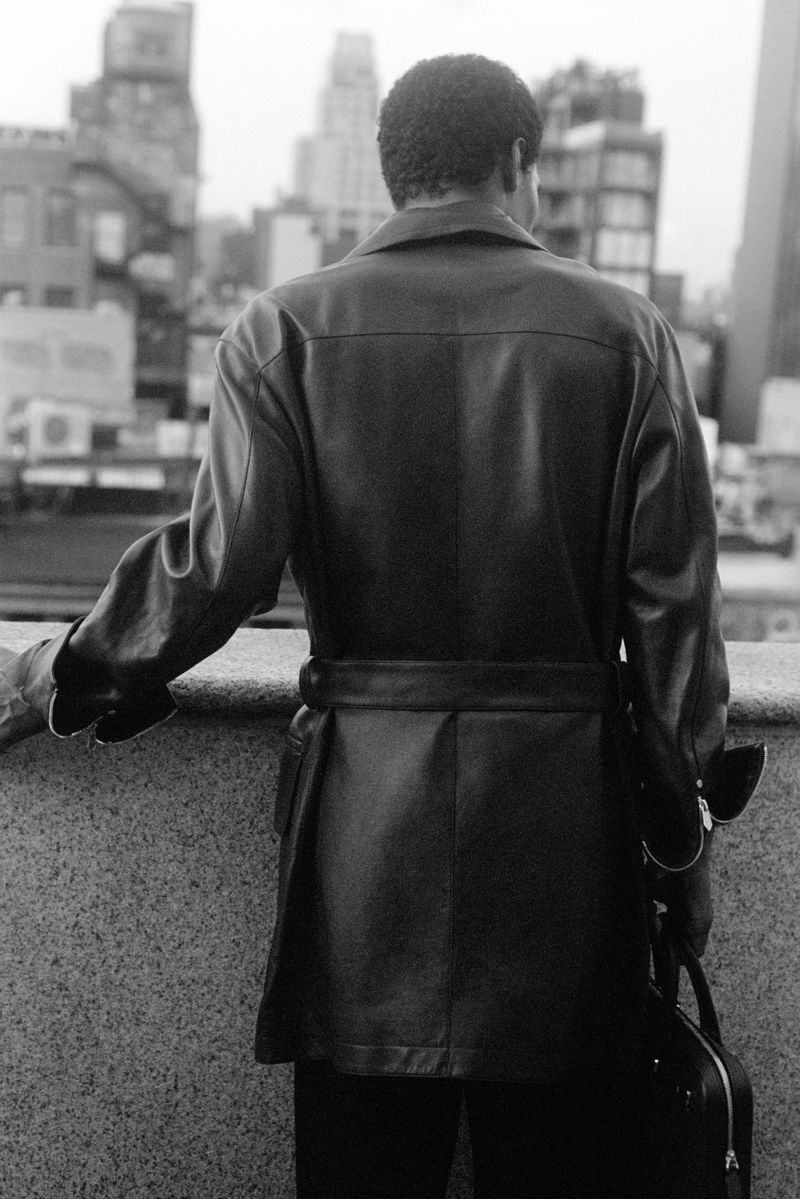 Back view of a man in a leather jacket and dark pants, holding a bag in a city setting with buildings in the background, black and white.