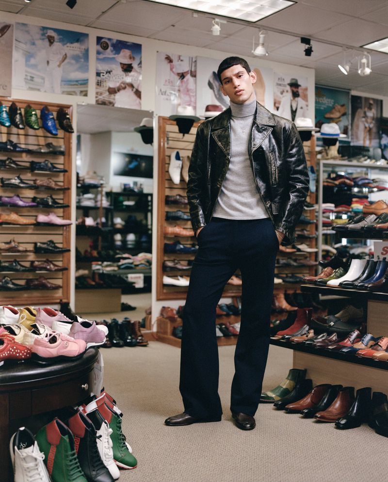 Stylish man in a leather jacket and trousers poses in the middle of a shoe store with shelves full of shoes and posters in the background.