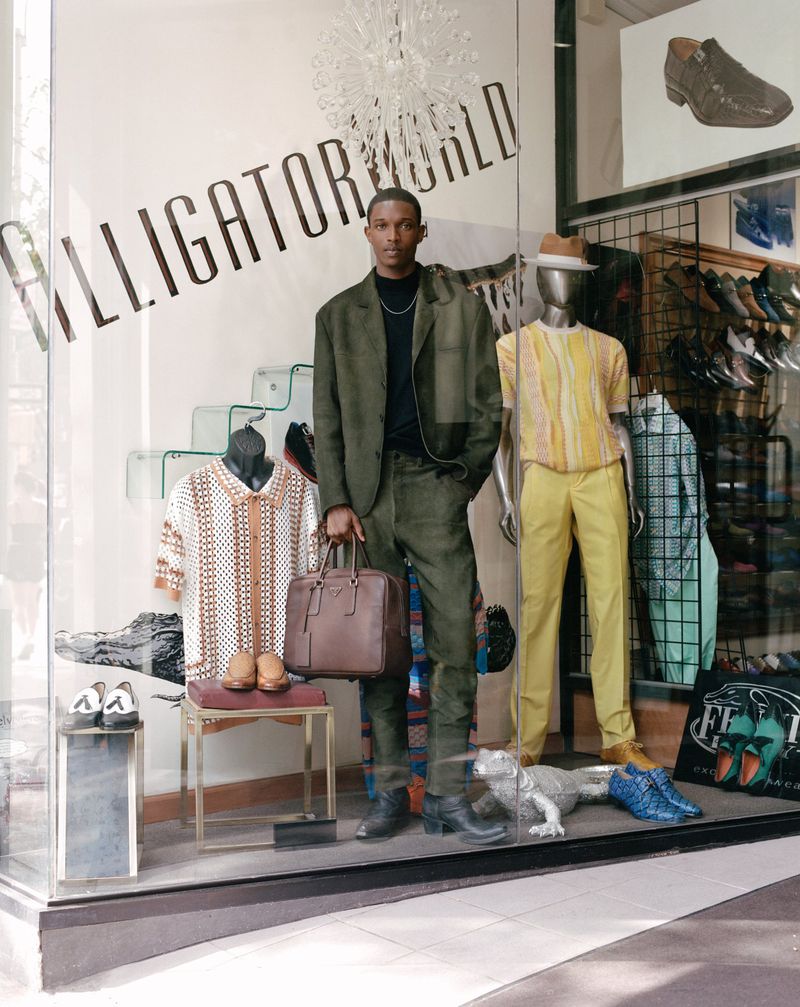 An adult male in a suit holds a briefcase in front of an Alligator World store window display with mannequins and shoes.