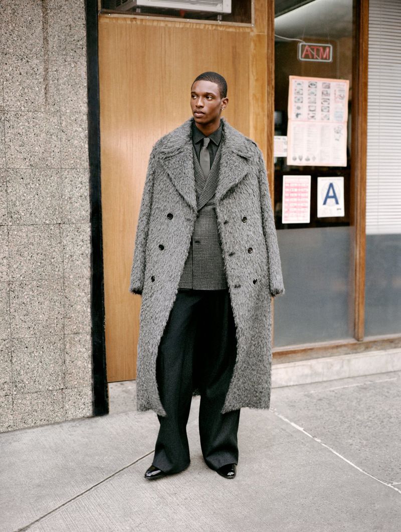 An African American man stands on a city sidewalk. He wears a gray double-breasted overcoat, suit, tie, black trousers, and black patent leather shoes.