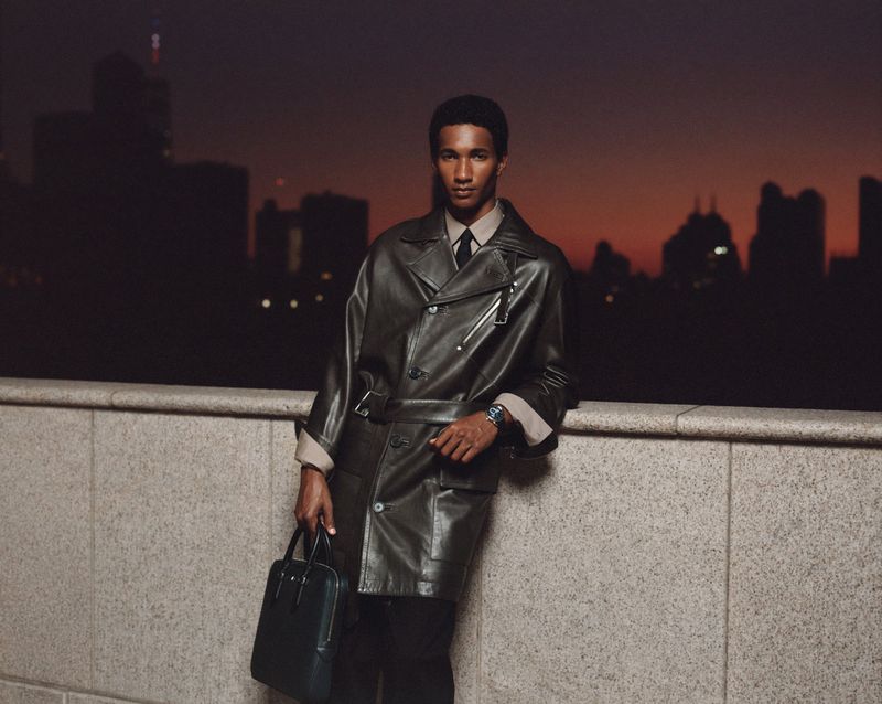 Fashion shot of a man leaning against a wall at dusk in a leather coat and carrying a briefcase, with a city skyline in the background.