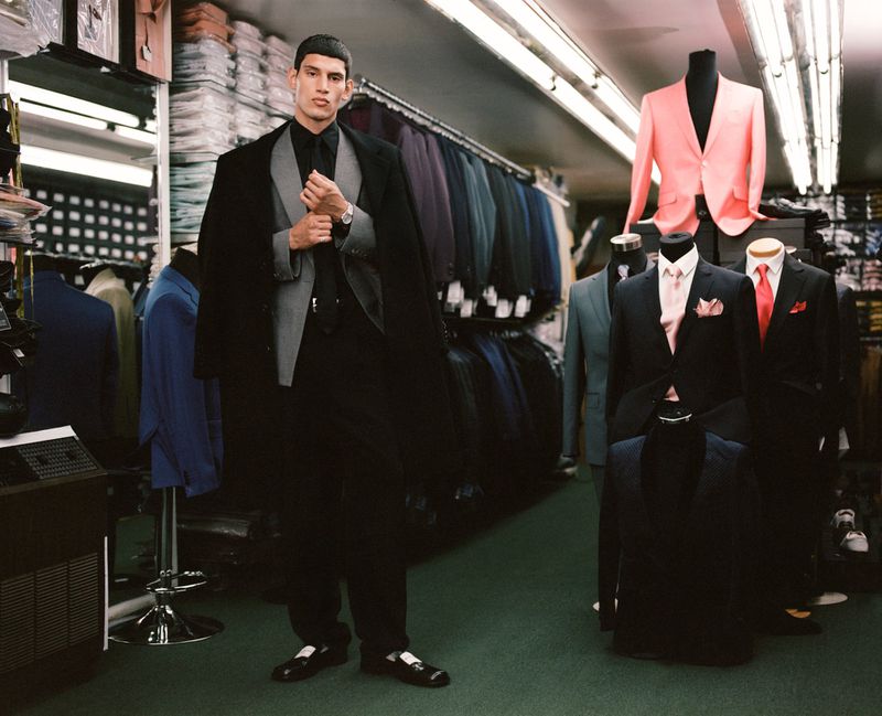 Man in black suit posing in tailored clothing store with formal wear. Mannequins showcase a variety of jackets and suits on display.