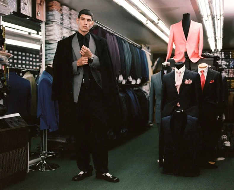 Man in black suit posing in tailored clothing store with formal wear. Mannequins showcase a variety of jackets and suits on display.