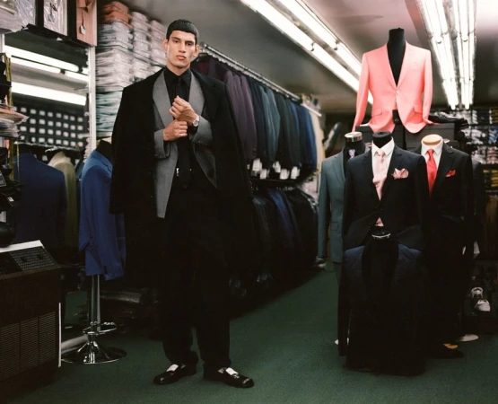 Man in black suit posing in tailored clothing store with formal wear. Mannequins showcase a variety of jackets and suits on display.