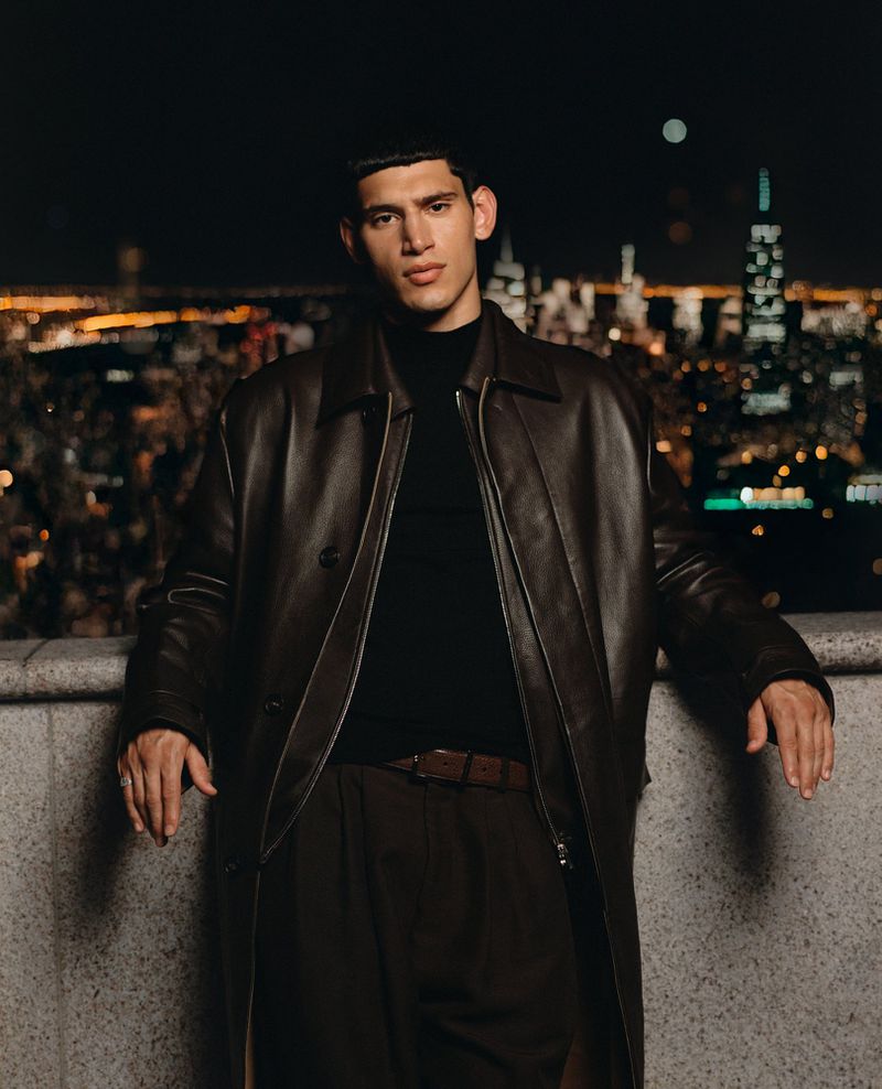 Handsome man poses in brown leather coat against a backdrop of city lights at night, creating a striking fashion portrait.