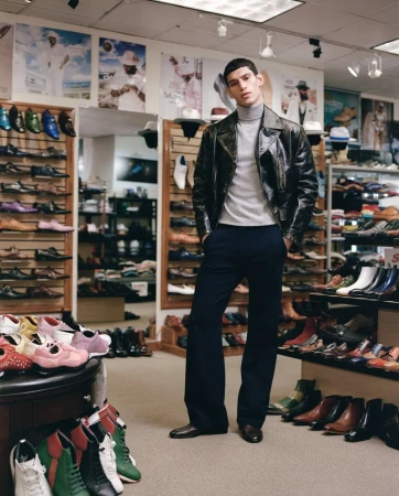 Model wearing a dark leather jacket and grey turtleneck standing inside a retail shoe store