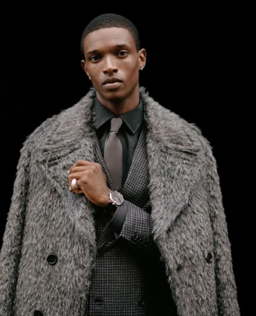 Close-up portrait of a Black man wearing a textured grey faux-fur coat, dark shirt, tie, and patterned waistcoat against a black background.